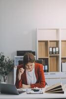 mujeres asiáticas sentadas en una oficina en casa con estrés y tensión ocular. mujer de negocios cansada sosteniendo anteojos y masajeando el puente nasal. hay tabletas, computadoras portátiles y café. foto