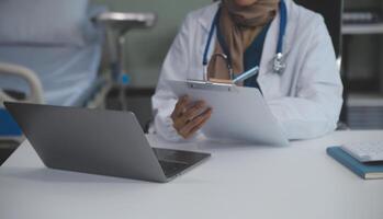 A professional and focused Asian female doctor in scrubs is working and reading medical research on her laptop in her office at a hospital. photo