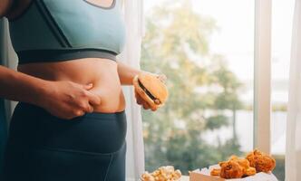 Portrait of obese woman looks sad while pinching her belly fat surrounded by junk foods. Diet fail concept photo