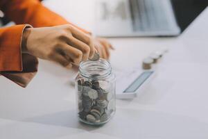 hermosa asiático mujer de negocios insertar monedas en un alfiler cerdito banco en el mesa. ahorro dinero, inversión, moneda concepto foto