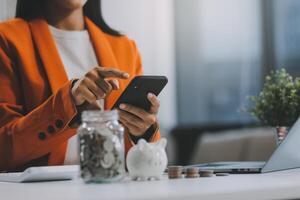 hermosa asiático mujer de negocios insertar monedas en un alfiler cerdito banco en el mesa. ahorro dinero, inversión, moneda concepto foto