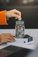 hermosa asiático mujer de negocios insertar monedas en un alfiler cerdito banco en el mesa. ahorro dinero, inversión, moneda concepto foto