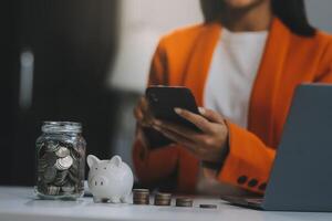 hermosa asiático mujer de negocios insertar monedas en un alfiler cerdito banco en el mesa. ahorro dinero, inversión, moneda concepto foto