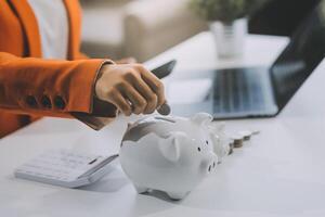 Beautiful Asian businesswoman insert coins in a pin piggy bank on the table. saving money, investment, currency concept photo