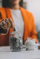 hermosa asiático mujer de negocios insertar monedas en un alfiler cerdito banco en el mesa. ahorro dinero, inversión, moneda concepto foto