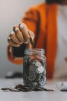 hermosa asiático mujer de negocios insertar monedas en un alfiler cerdito banco en el mesa. ahorro dinero, inversión, moneda concepto foto