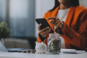 hermosa asiático mujer de negocios insertar monedas en un alfiler cerdito banco en el mesa. ahorro dinero, inversión, moneda concepto foto