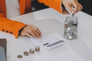 hermosa asiático mujer de negocios insertar monedas en un alfiler cerdito banco en el mesa. ahorro dinero, inversión, moneda concepto foto