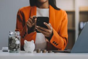 Beautiful Asian businesswoman insert coins in a pin piggy bank on the table. saving money, investment, currency concept photo