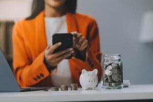 hermosa asiático mujer de negocios insertar monedas en un alfiler cerdito banco en el mesa. ahorro dinero, inversión, moneda concepto foto