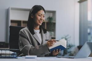 Confident millennial indian female business analyst financial advisor preparing statistic report studying documents on work desk, browsing information online using pc, writing out notes on paper sheet photo