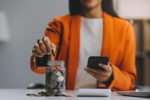 hermosa asiático mujer de negocios insertar monedas en un alfiler cerdito banco en el mesa. ahorro dinero, inversión, moneda concepto foto