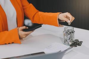 hermosa asiático mujer de negocios insertar monedas en un alfiler cerdito banco en el mesa. ahorro dinero, inversión, moneda concepto foto