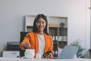 hermosa asiático mujer de negocios insertar monedas en un alfiler cerdito banco en el mesa. ahorro dinero, inversión, moneda concepto foto