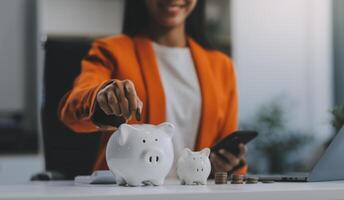 Beautiful Asian businesswoman insert coins in a pin piggy bank on the table. saving money, investment, currency concept photo