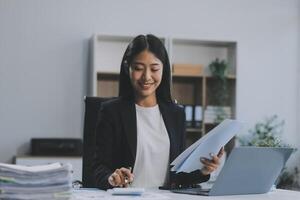 Confident millennial indian female business analyst financial advisor preparing statistic report studying documents on work desk, browsing information online using pc, writing out notes on paper sheet photo