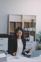 Confident millennial indian female business analyst financial advisor preparing statistic report studying documents on work desk, browsing information online using pc, writing out notes on paper sheet photo