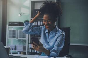 Happy excited successful Asian businesswoman triumphing with a laptop computer smartphone in the workplace office photo