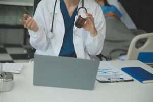 un hermosa y profesional asiático hembra médico es trabajando en su médico casos en su ordenador portátil o haciendo un llamada médico consulta con un paciente mientras sentado en su oficina. foto