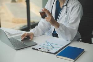 un hermosa y profesional asiático hembra médico es trabajando en su médico casos en su ordenador portátil o haciendo un llamada médico consulta con un paciente mientras sentado en su oficina. foto