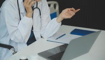 un hermosa y profesional asiático hembra médico es trabajando en su médico casos en su ordenador portátil o haciendo un llamada médico consulta con un paciente mientras sentado en su oficina. foto