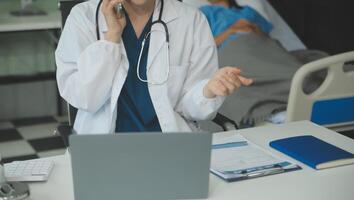 un hermosa y profesional asiático hembra médico es trabajando en su médico casos en su ordenador portátil o haciendo un llamada médico consulta con un paciente mientras sentado en su oficina. foto