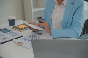 encantadora joven mujer de negocios asiática sentada en una computadora portátil en la oficina, haciendo un informe que calcula el documento de verificación del servicio de ingresos internos del saldo. foto