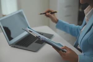 encantadora joven mujer de negocios asiática sentada en una computadora portátil en la oficina, haciendo un informe que calcula el documento de verificación del servicio de ingresos internos del saldo. foto