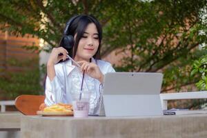 oficina hermosa asiático mujer en al aire libre cafetería. niña con un agraciado sonrisa escuchando a música mientras participación un bolígrafo haciendo trabajo en frente de un ordenador portátil para contenido o promociones foto