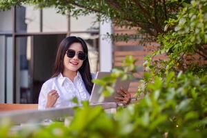 oficina hermosa asiático mujer en al aire libre cafetería. alegre niña mirando a el cámara exitosamente completa el trabajo en el ordenador portátil participación un bolígrafo vistiendo Gafas de sol para contenido o publicidad foto