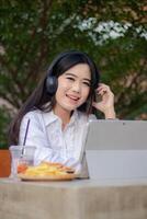 office beautiful asian woman in outdoor cafe. smiling girl laughing listening to music while typing doing work in front of the laptop looking at the camera for content or advertisements photo