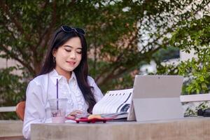 oficina hermosa asiático mujer en al aire libre cafetería. sonriente niña comprobación trabajo mientras leyendo un libro vistiendo Gafas de sol para contenido o publicidad foto