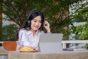 office beautiful asian woman in outdoor cafe. spoiled smiling girl listening to music while typing doing work in front of the laptop looking at the front for content or promos photo