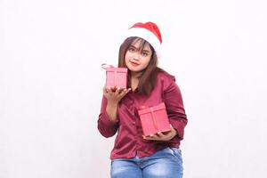 beautiful young asian indonesian girl carrying gift box at christmas santa claus hat modern shiny red shirt outfit looking at camera plain on white background for promotion and advertising photo