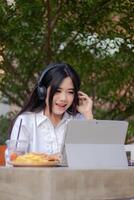 office beautiful asian woman in outdoor cafe. spoiled smiling girl listening to music while typing doing work in front of a laptop for content or promos photo