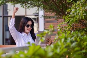 office beautiful asian woman in outdoor cafe. cheerful candid girl successfully completes work on laptop holding pen wearing sunglasses for content or advertising photo