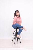 beautiful cheerful black-haired woman in her 20s wearing a basic pink shirt working sitting in a chair with crossed legs studio portrait on white background for service promotion photo