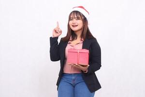 Beautiful Asian girl in cheerful suit with Santa Claus hat posing with Christmas gift box and hands pointing up on white background for promotion, advertising, banner, billboard full photo