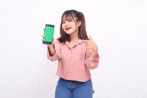 Beautiful happy Asian woman in her 20s wearing casual shirt holding green screen cell phone laughing pointing at gadget on white background studio portrait for banner ad, banner, billboard photo