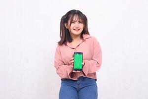 hermosa contento asiático mujer en su 20s vistiendo casual camisa participación verde pantalla Teléfono móvil mientras sonriente descuidadamente en blanco antecedentes estudio retrato para bandera anuncio, bandera, cartelera foto