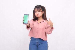 Beautiful happy Asian woman in her 20s wearing casual shirt holding white screen cellphone while frowning pointing at gadget on white background studio portrait for banner ad, banner, billboard photo