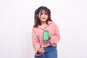 Beautiful happy asian woman in her 20s wearing casual shirt holding cellphone green screen pointing gadget from below on white background studio portrait for banner ad, banner, billboard photo