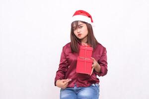 beautiful young asian indonesian girl pouting carrying gift box in christmas santa claus hat outfit red shirt modern pointed bottom box on white background for promotion and advertising photo
