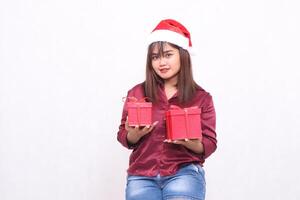 Photo portrait of beautiful young asian girl carrying gift box in christmas santa claus hat modern shiny red shirt outfit hands raise box and front view on white background for promotion
