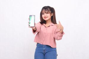 hermosa contento asiático mujer en su 20s vistiendo casual camisa participación blanco pantalla Teléfono móvil mientras sonriente señalando a artilugio en blanco antecedentes estudio retrato para bandera anuncio, bandera, cartelera foto