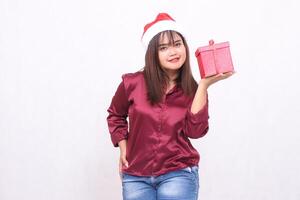 Photo portrait of beautiful asian girl kissing age carrying gift box in christmas santa claus hat modern shiny red shirt outfit left hand lifting box on white background for promotion and advertising