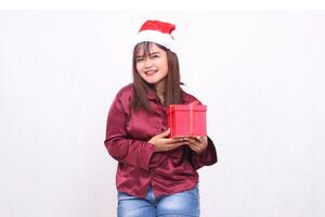 Photo portrait of beautiful cheerful Asian girl in her 20s carrying boxed gifts in Christmas Santa Claus hat wearing shiny modern red shirt seen on white background for promotion and advertising
