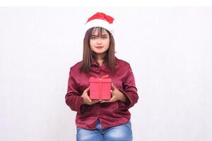 foto retrato de un hermosa llanura asiático niña en su 20s que lleva un regalo caja a Navidad Papa Noel claus sombrero vistiendo un moderno brillante rojo camisa visto en un blanco antecedentes para promoción y publicidad