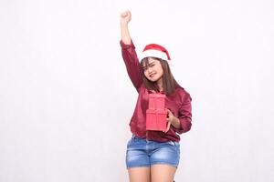 Cheerful young beautiful Southeast Asian girl carrying gift box at Christmas wearing Santa Claus hat modern red shirt outfit raising hands on white background for promotion and advertising photo
