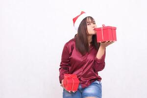 Photo portrait of young beautiful asian girl carrying gift box in christmas santa claus hat modern shiny red shirt outfit kissing box looking at camera on white background for promotion ,advertising